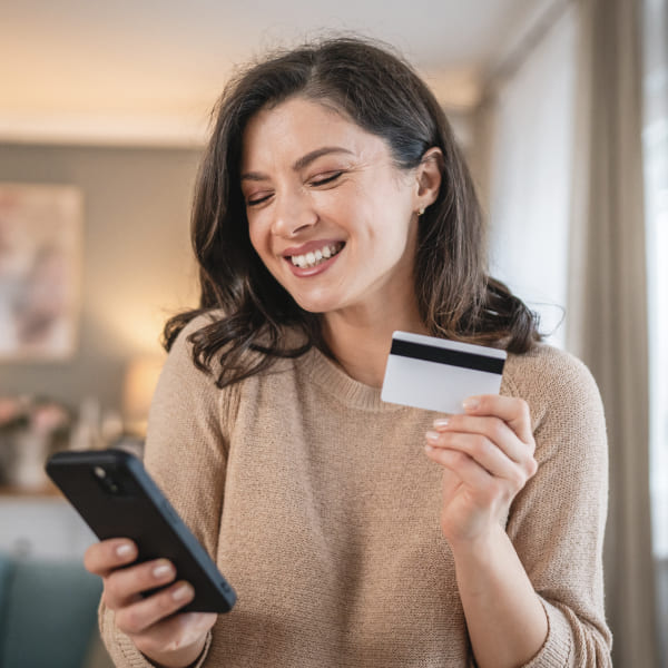 Femme souriante tenant une carte de crédit et utilisant son smartphone à la maison.